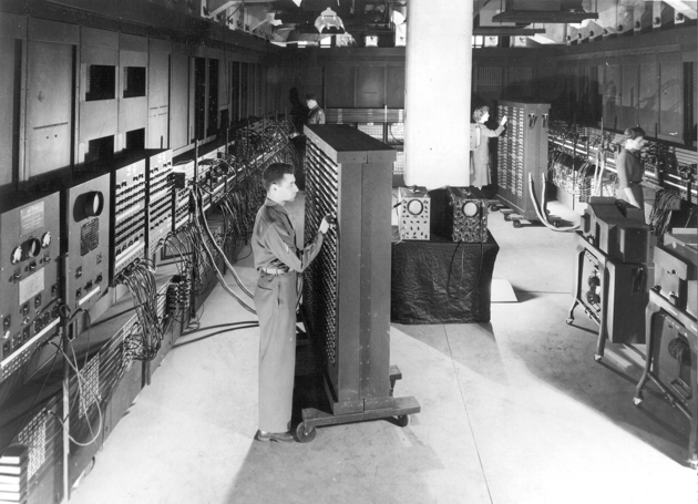 Room-sized ENIAC computer with rows of vacuum tubes, cables, and control panels from 1945
